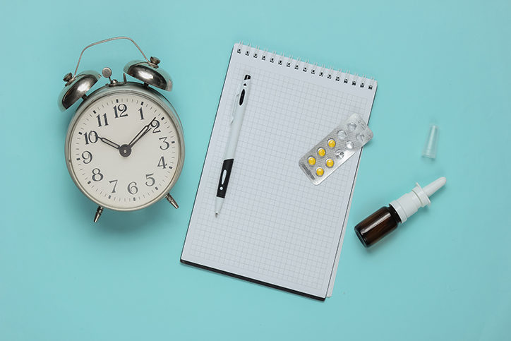 Medical still life on a blue studio background. Top view. Flat lay. Items include a clock, notepad, pill pack, and nasal spray.